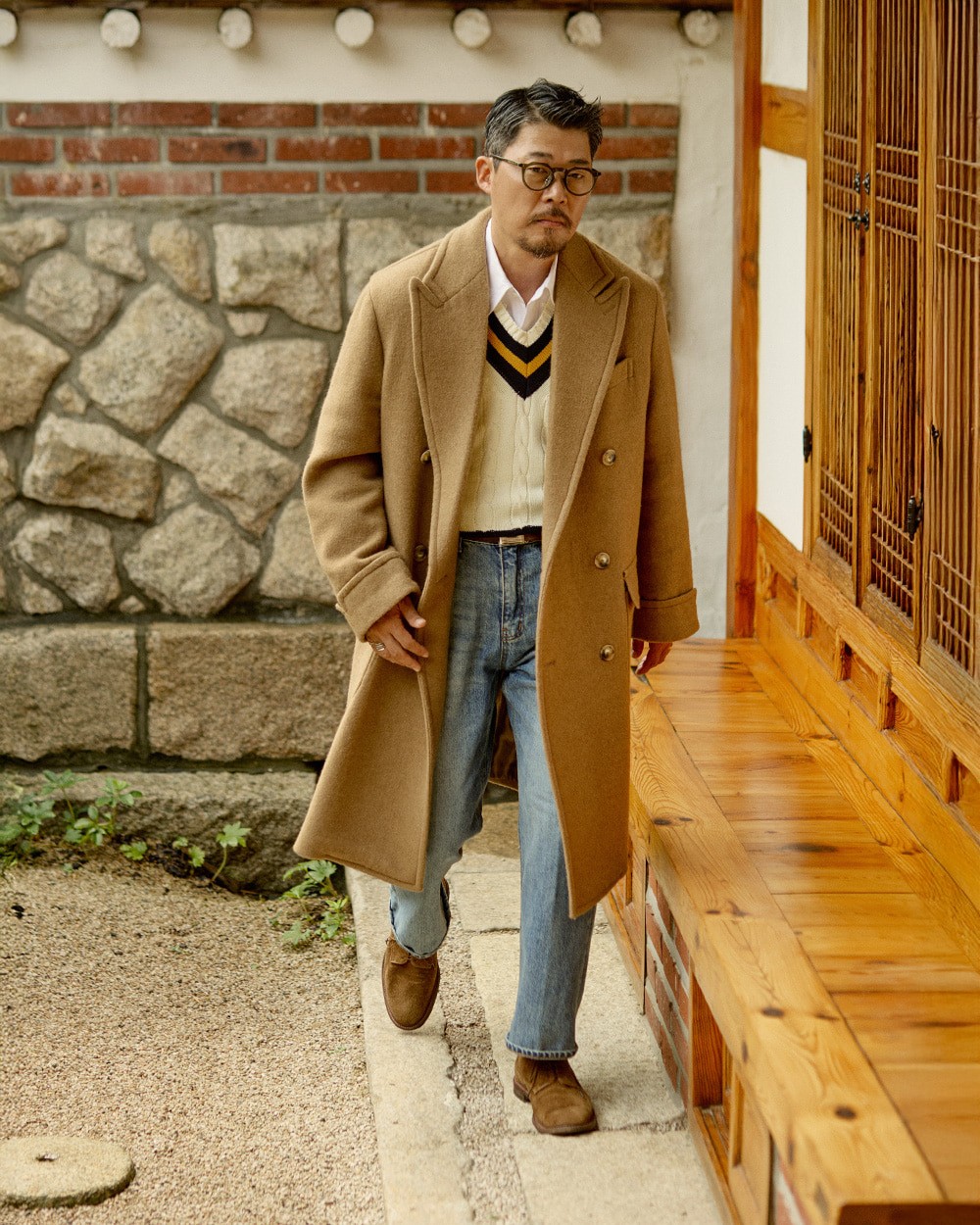 Man in a camel polo coat open over an ivory cricket sweater with navy stripes, with blue jeans and brown suede shoes, in a hanok courtyard