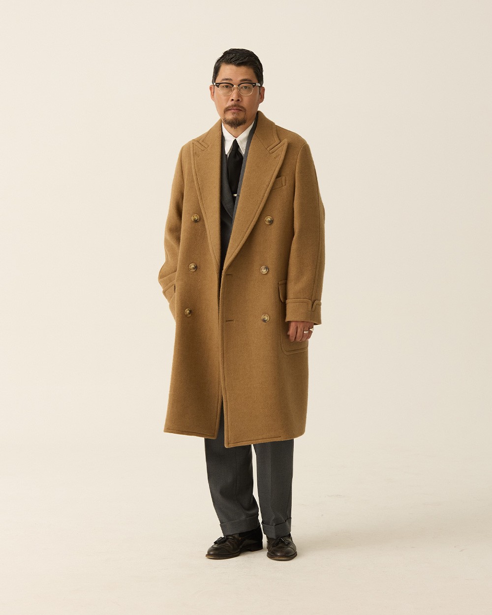 Man in a camel polo coat open over a navy jacket and dark tie, with grey trousers and black loafers