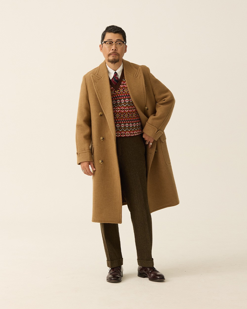 Man in a camel polo coat open over a red patterned Fair Isle vest, white shirt and striped tie, with brown herringbone trousers and burgundy loafers