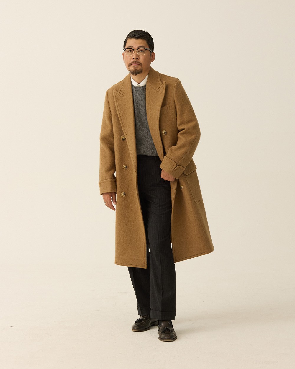 Man in a camel polo coat open over a grey crewneck sweater and white shirt collar, with charcoal trousers and black loafers