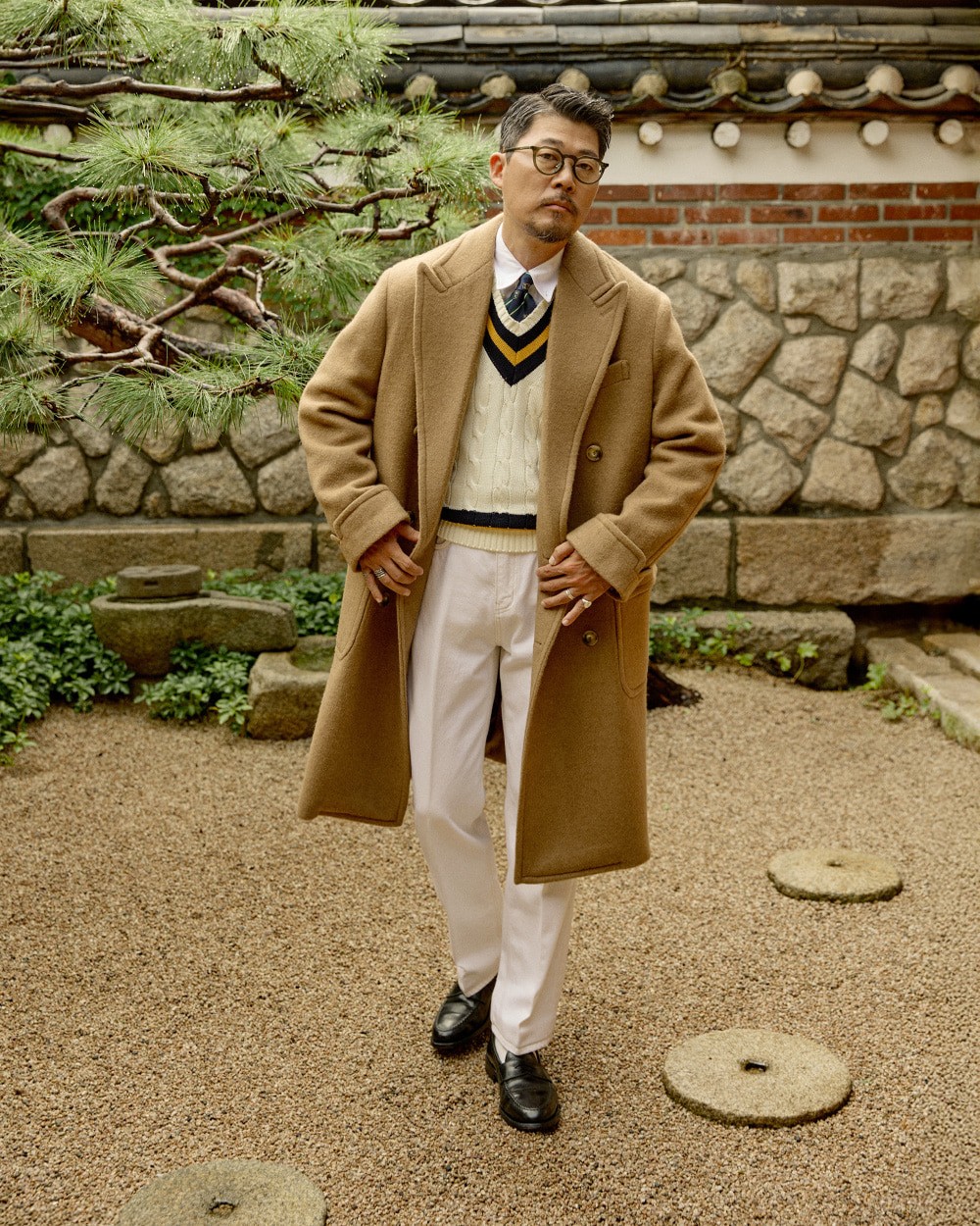Man in a camel polo coat open over an ivory cricket sweater with navy stripes, white shirt and tie, with cream trousers and black loafers, in a hanok garden