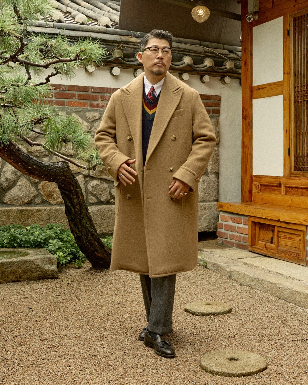 Man in a camel polo coat open over a navy cricket sweater with red and yellow stripes, white shirt and striped tie, with grey trousers and black loafers, under a pine tree in a hanok garden