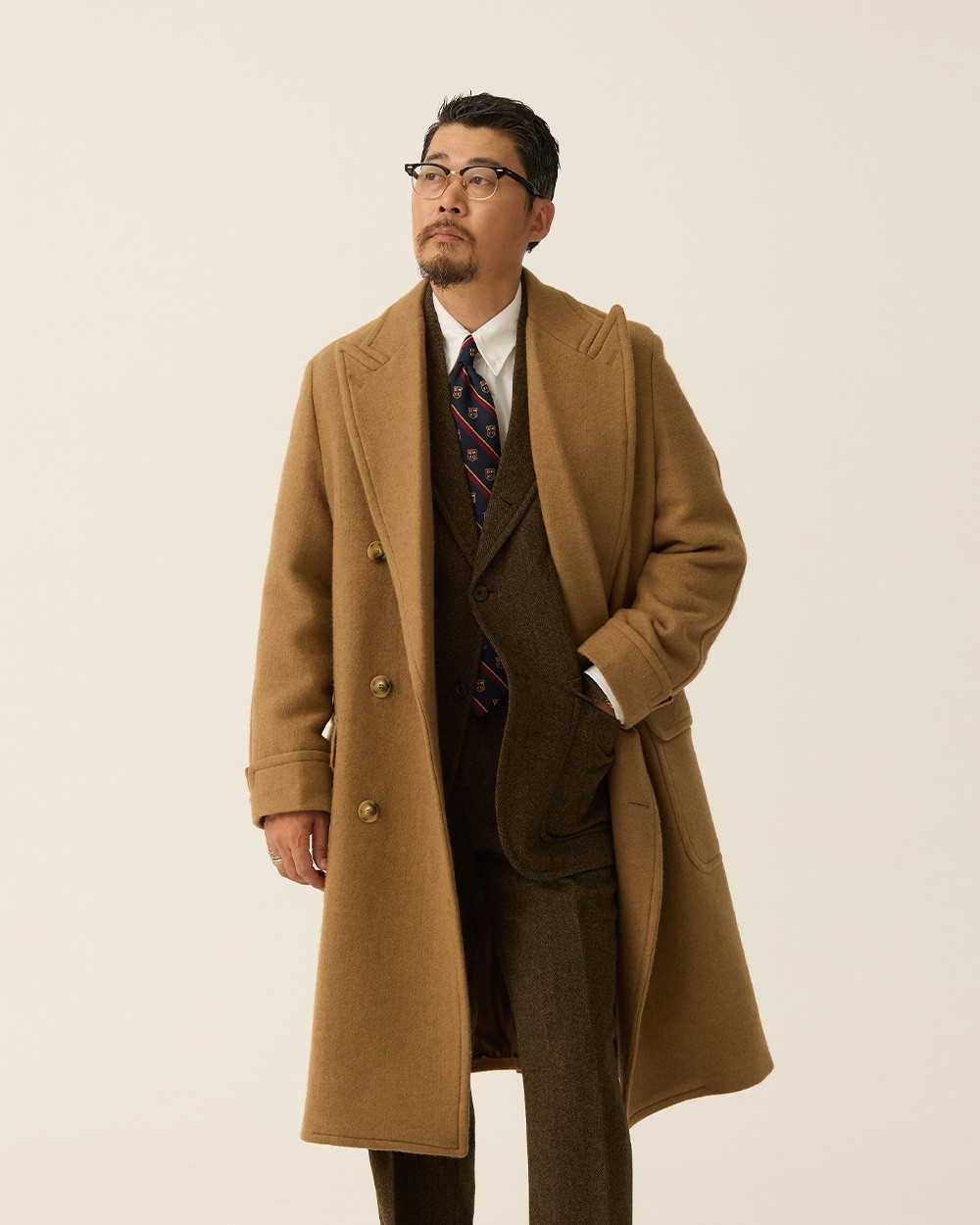 Close-up of a camel polo coat over an olive herringbone suit jacket and paisley tie, hands in the coat pockets