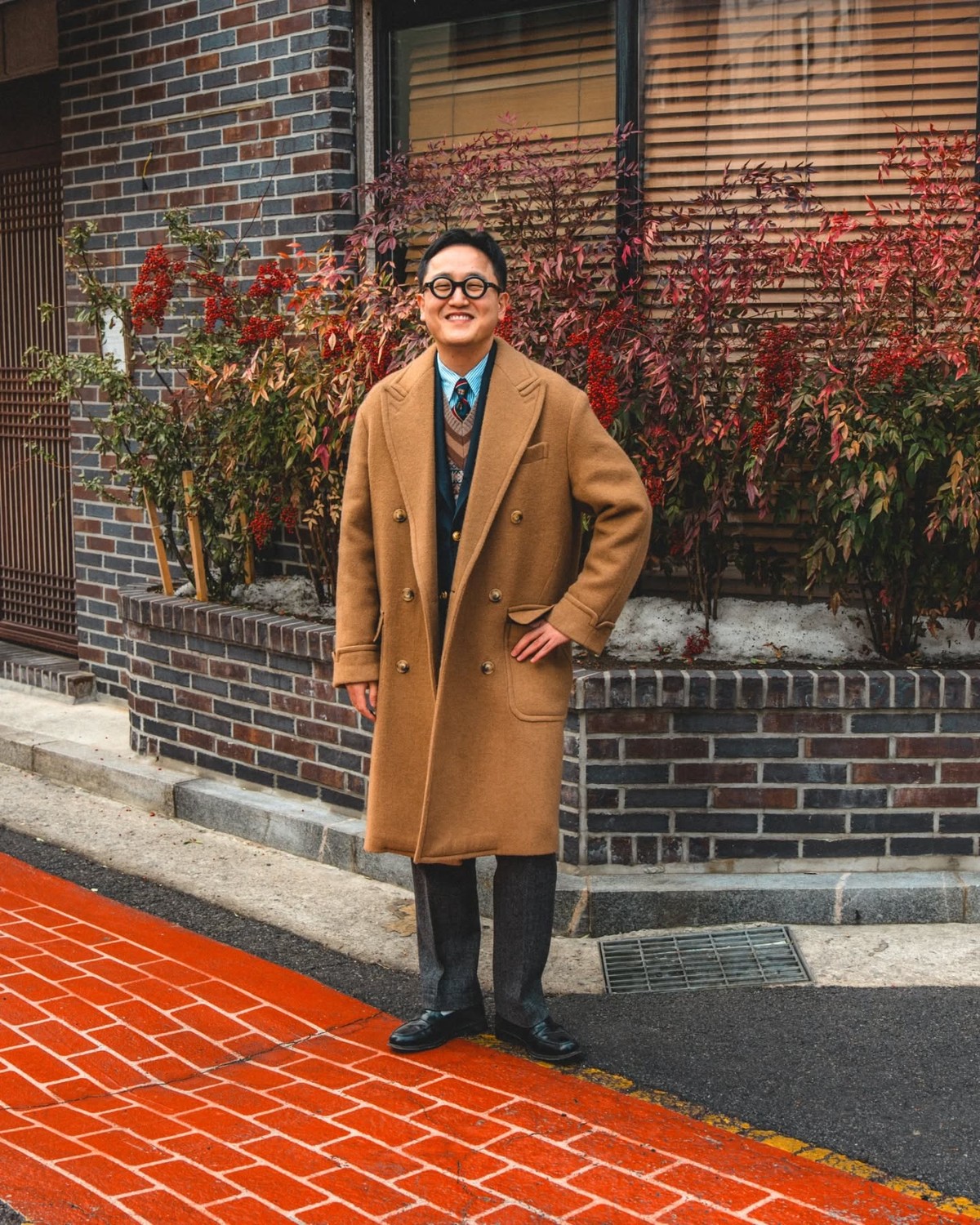 Man in a camel polo coat worn open over a navy jacket, a patterned V-neck knit, light blue shirt and striped tie, with grey flannel trousers and black loafers, in front of a brick wall with red leaves