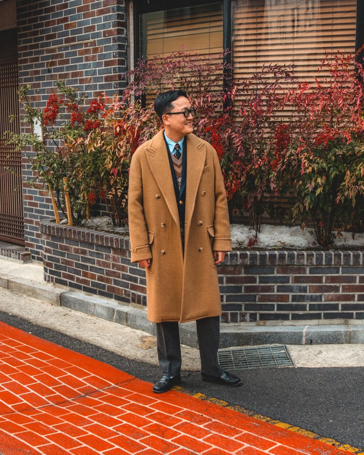 Man in a camel polo coat worn open over a navy jacket, patterned V-neck knit, light blue shirt and tie, with grey flannel trousers and black loafers, standing on a street beside a hedge with red leaves