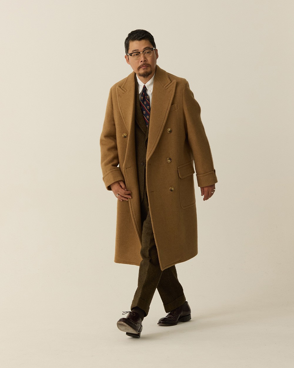 Man walking in a camel polo coat worn open over a brown suit and tie, with dark shoes