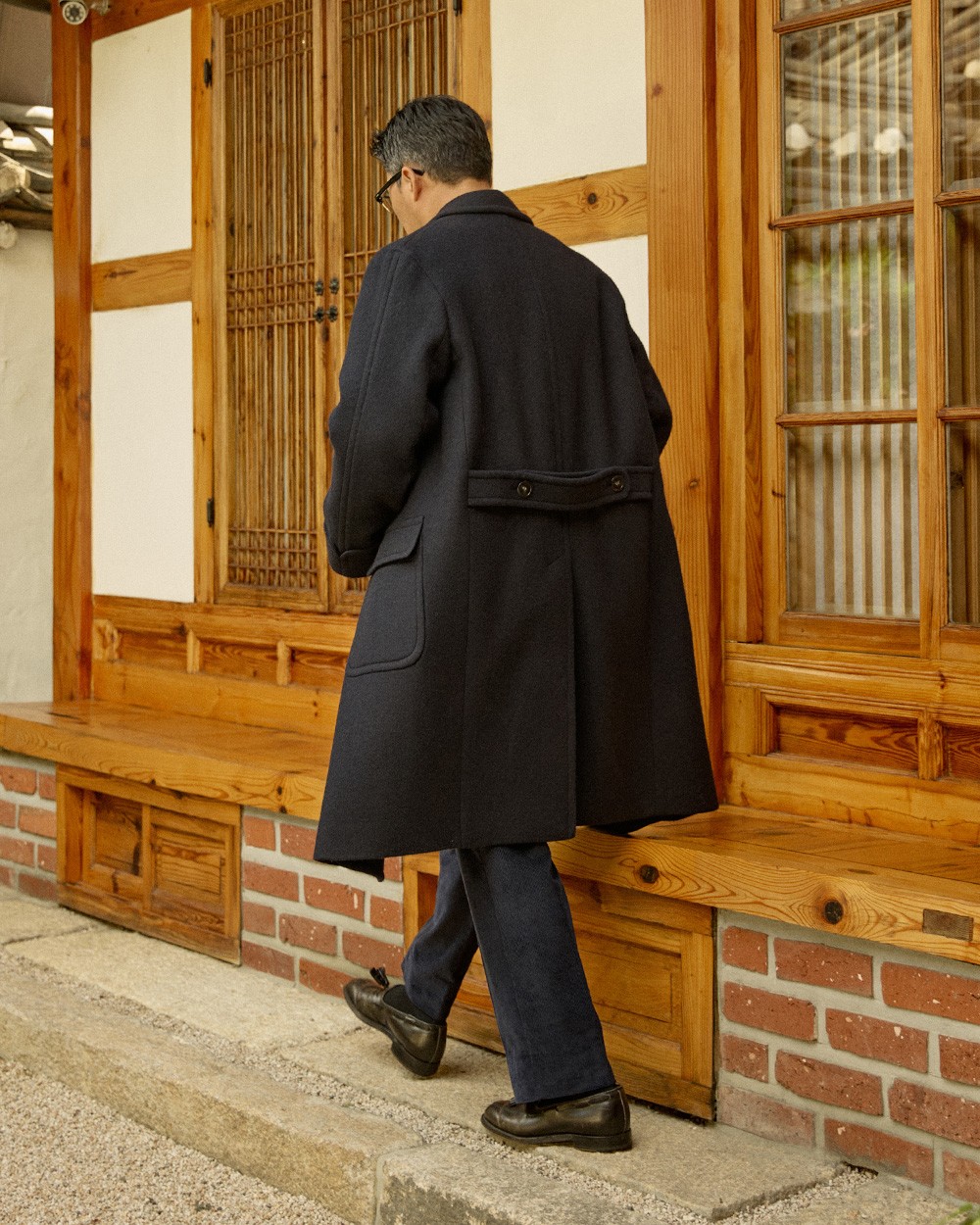 Back view of a man in a navy polo coat with dark jeans and black loafers, stepping onto a hanok's wooden floor, the half belt visible