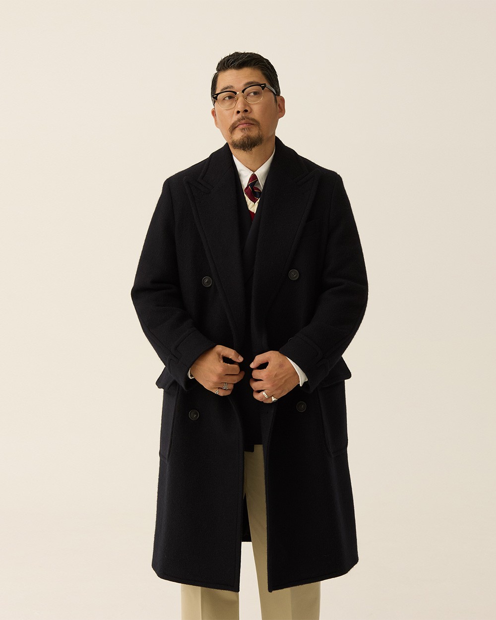 Man buttoning a navy polo coat over a navy jacket, white shirt and red-and-navy repp tie, with khaki trousers