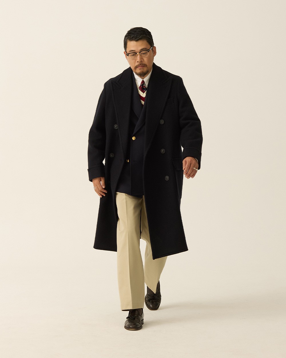 Man in a navy polo coat open over a navy gold-button blazer and striped tie, with khaki trousers and black loafers, the coat ending below the knee