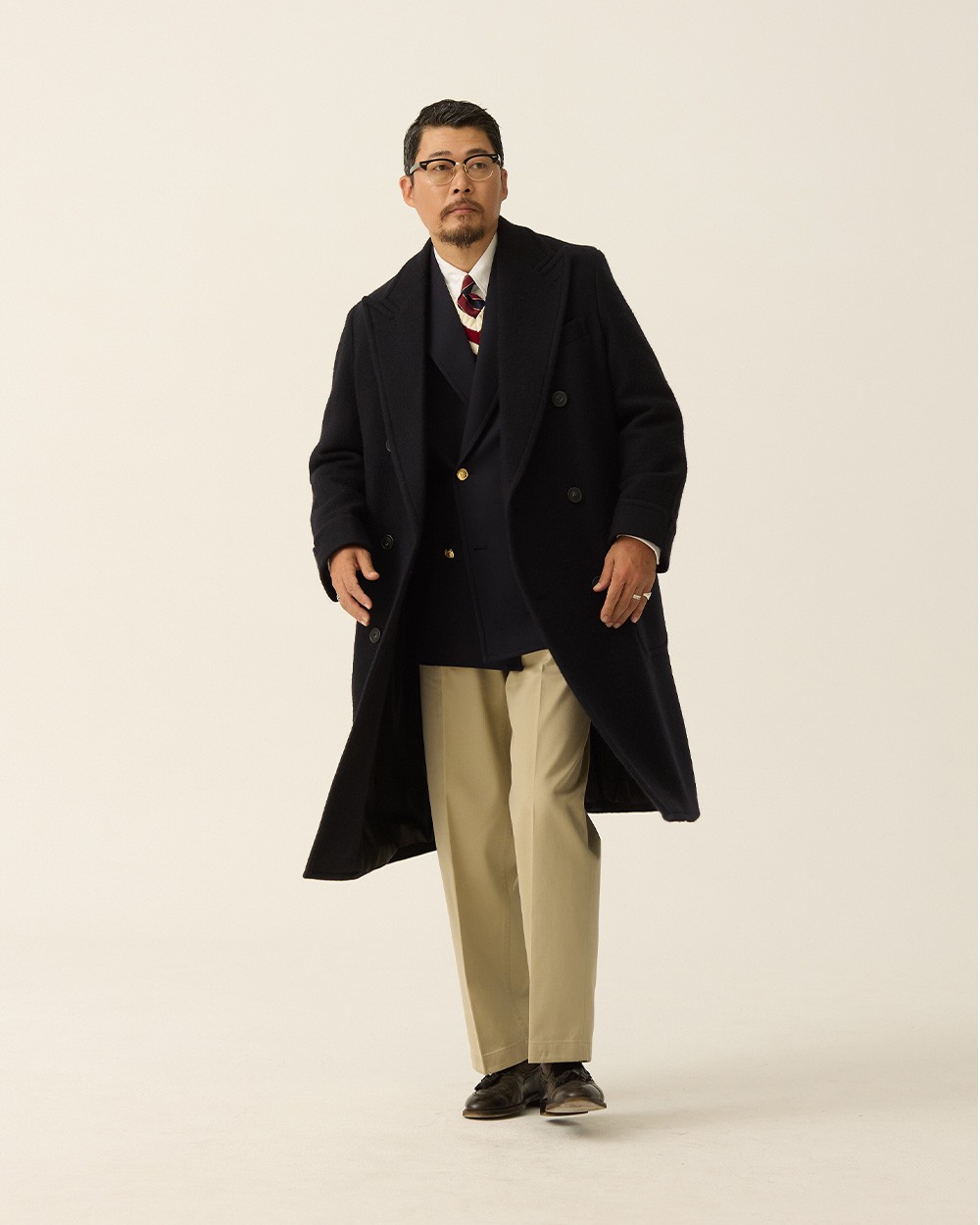 Man in a navy polo coat open over a navy gold-button blazer, white shirt and striped tie, with khaki trousers and black loafers