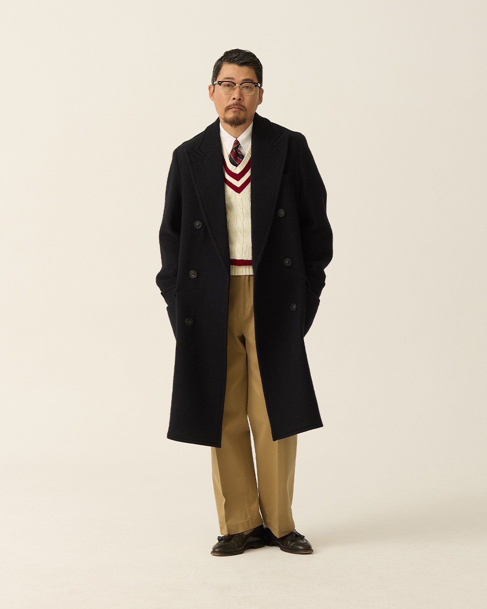 Man in a navy polo coat open over an ivory cricket sweater with red and navy stripes at the V-neck, with khaki trousers and brown loafers