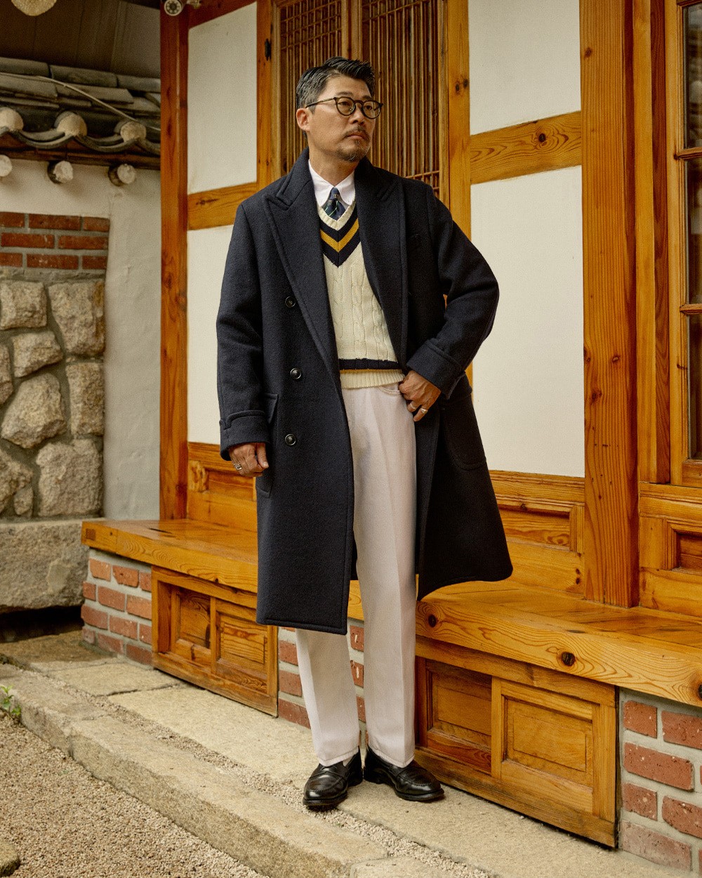Man in a navy polo coat open over an ivory cricket sweater with navy and yellow stripes, shirt and tie, with white trousers and black loafers, in front of a hanok
