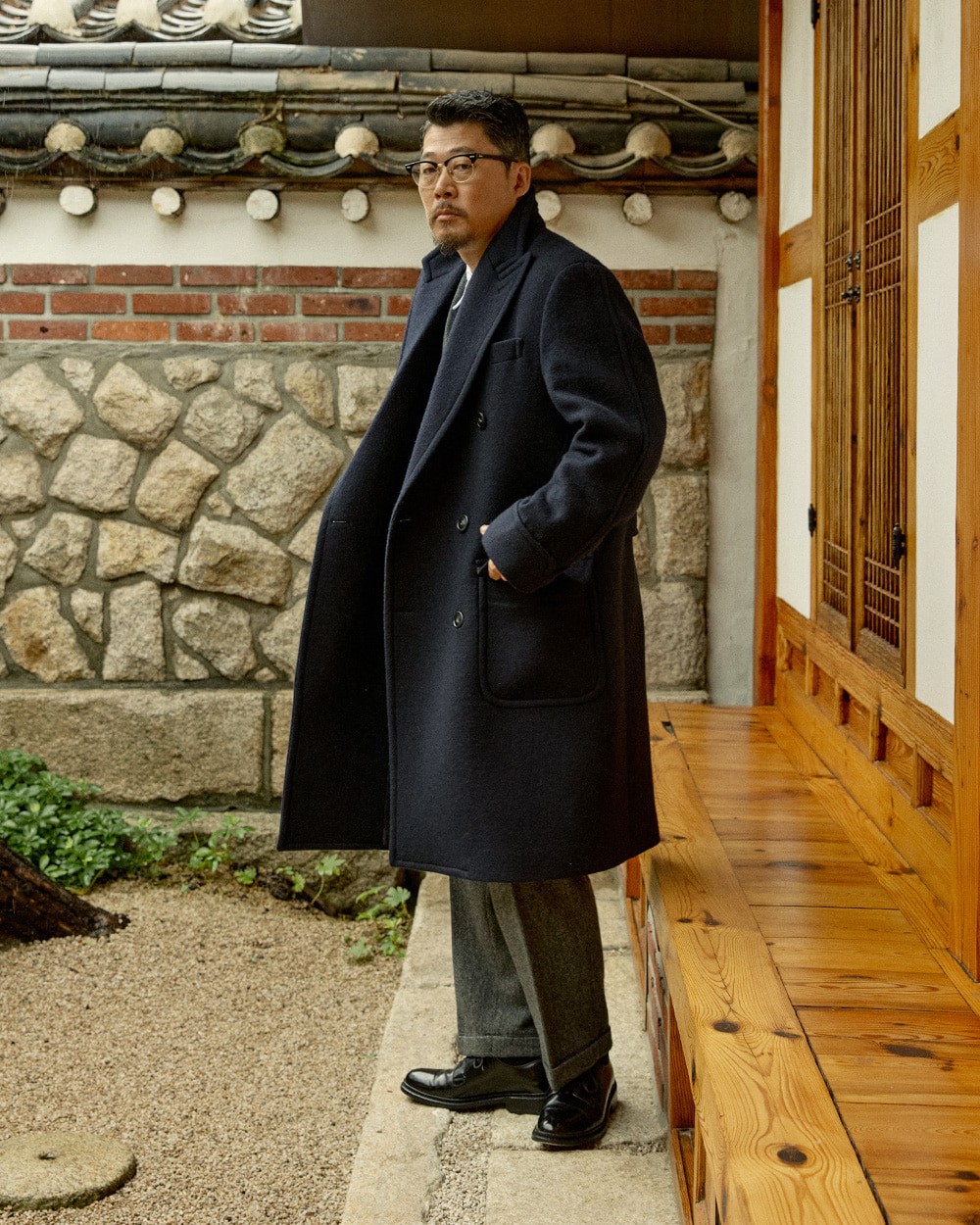 Man in a navy polo coat open over a navy crewneck sweater and white shirt collar, with grey trousers and black loafers, in a hanok courtyard