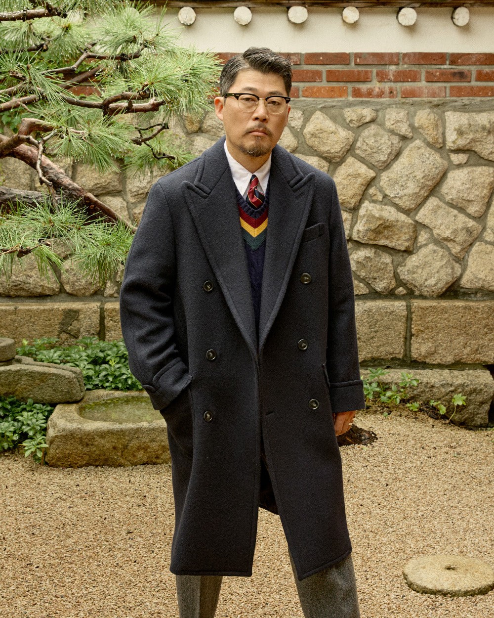 Close-up of a man in a navy polo coat over a navy cricket sweater with red, yellow and green stripes, white shirt and red-and-navy striped tie, in front of a pine tree