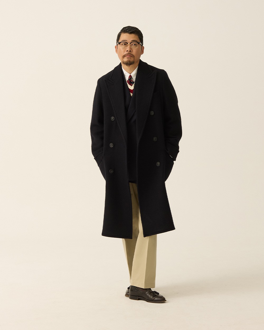 Man in a navy polo coat open over a navy jacket and tie, with khaki trousers and brown loafers