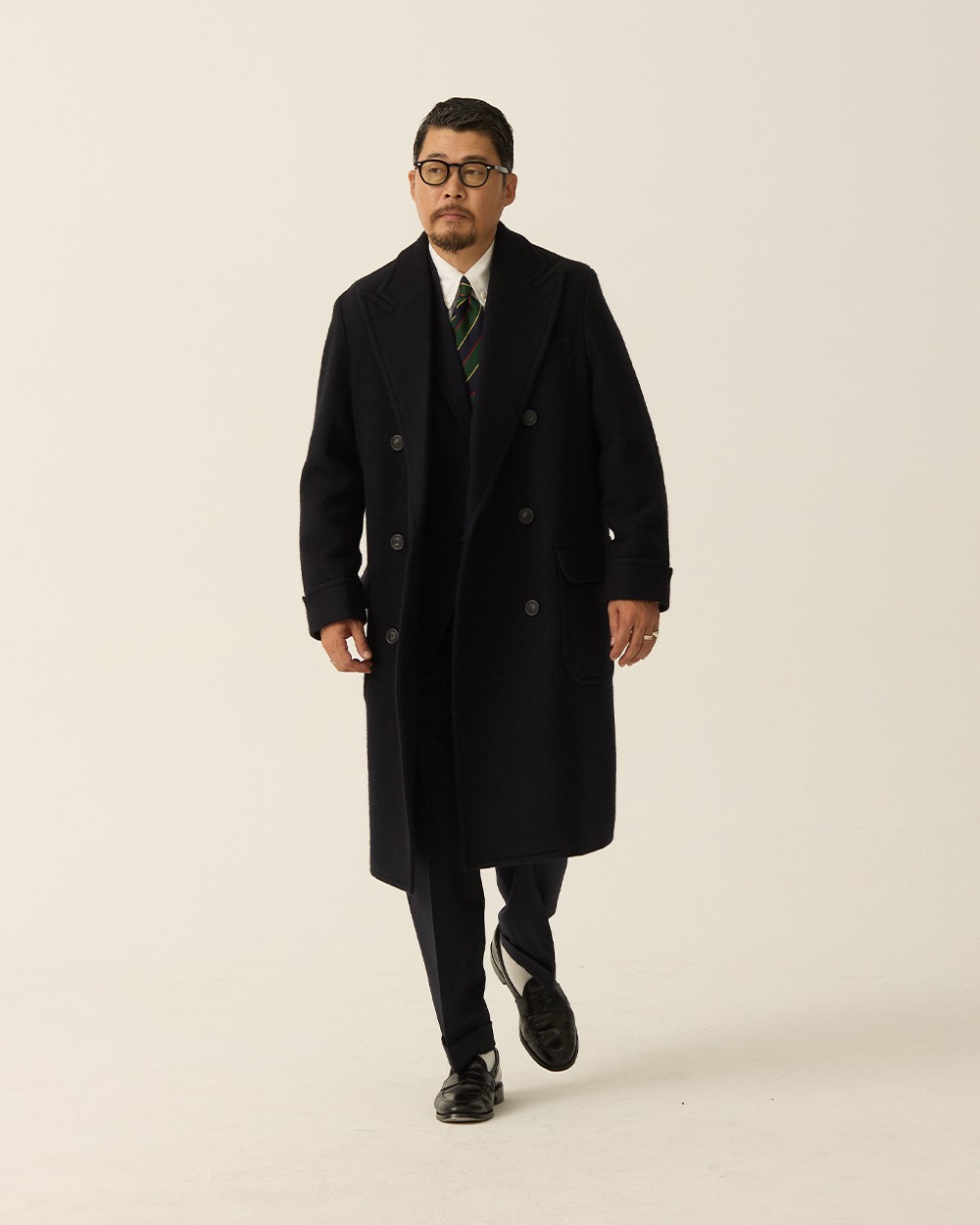 Man walking in a navy polo coat open over a navy suit, white shirt and green striped tie, with black loafers
