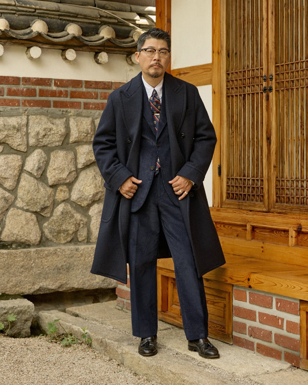 Man in a navy polo coat over a navy suit and striped tie, with black loafers, standing by a hanok stone wall