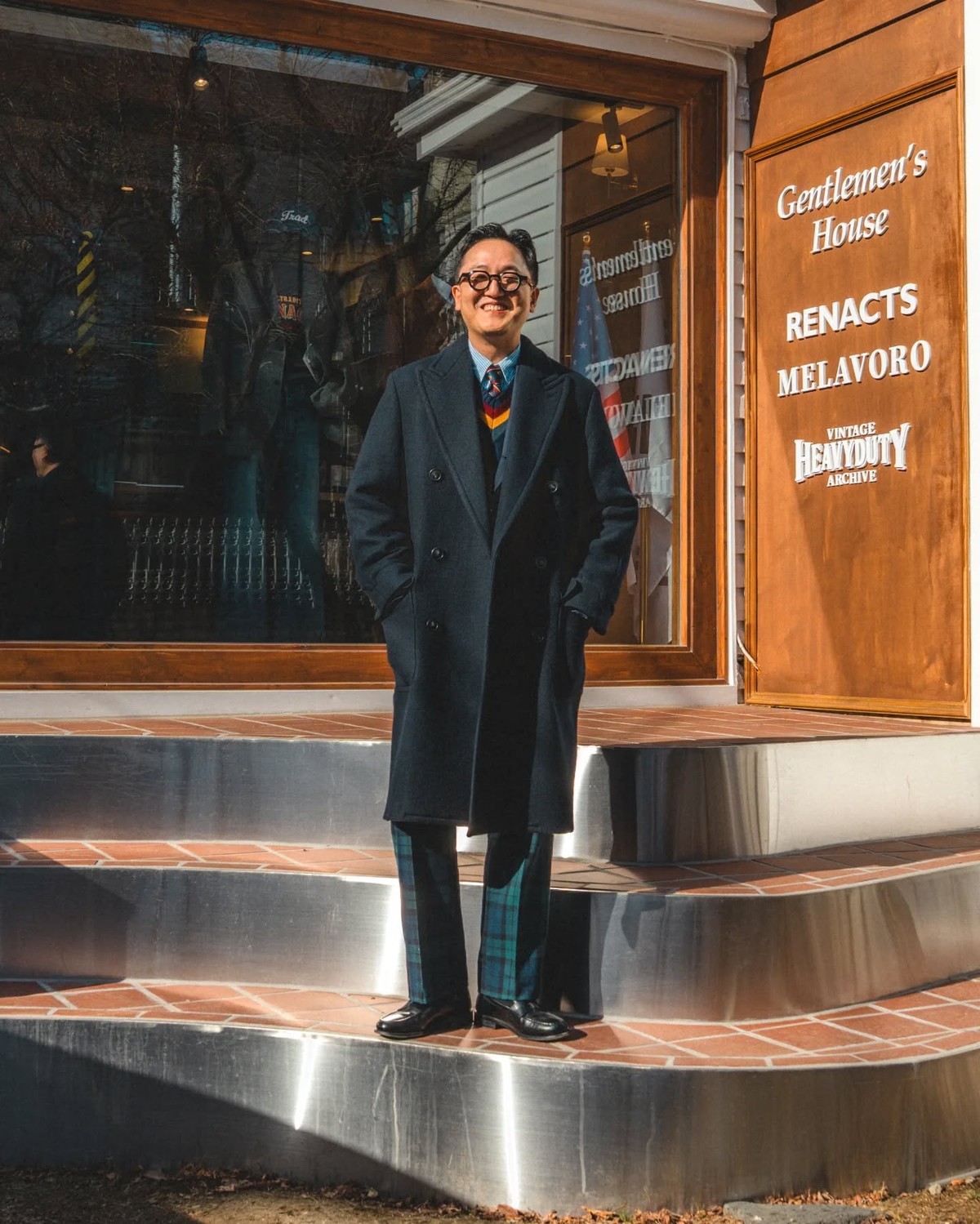 Man in a navy polo coat over a navy cricket sweater, light blue shirt and tie, with Black Watch tartan trousers and black loafers, standing on the metal step of a shop entrance and looking to the side