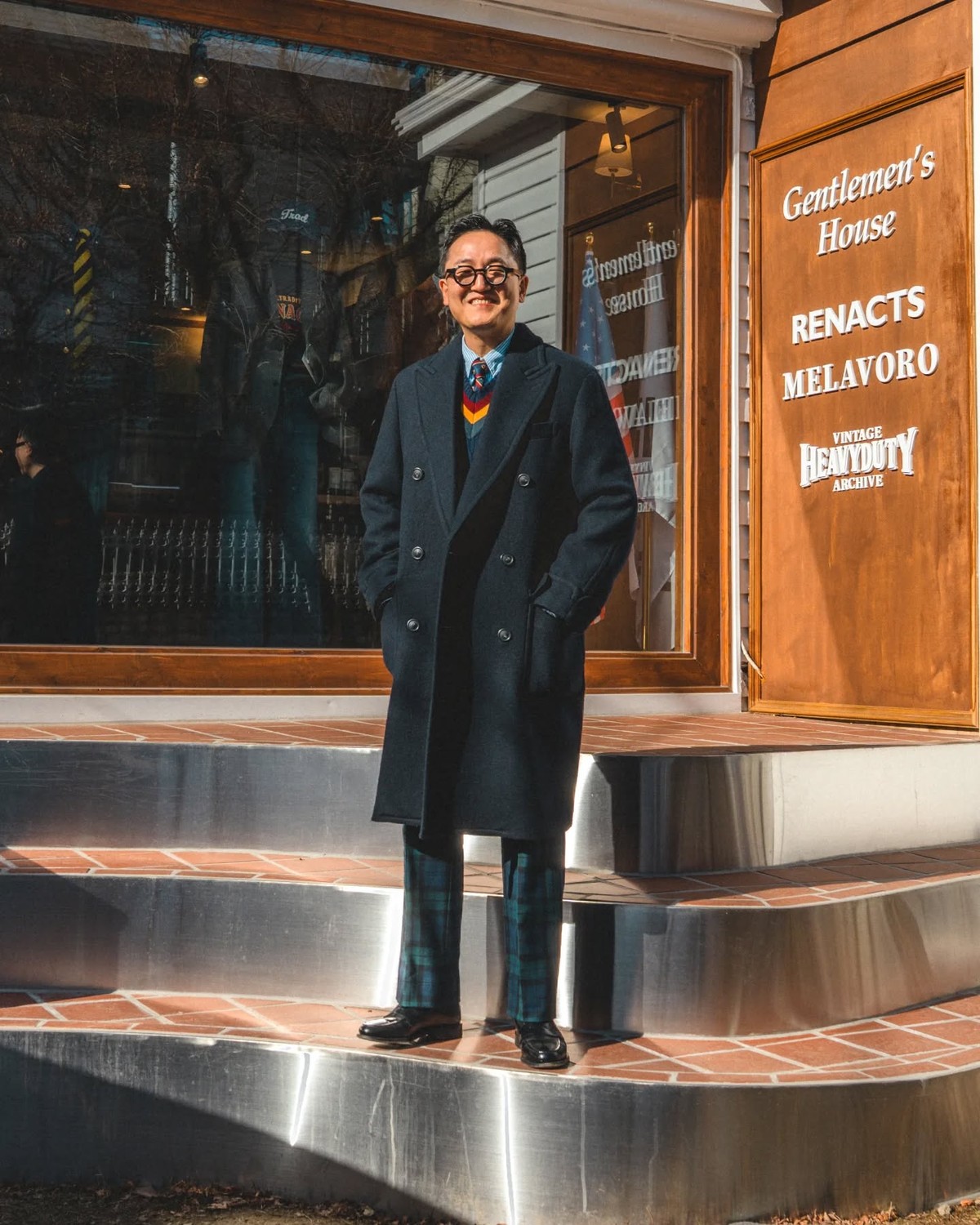 Man in a navy polo coat worn open over a navy cricket sweater with red, yellow and green stripes, light blue shirt and striped tie, with Black Watch tartan trousers and black loafers, in front of the Gentlemen's House shop window in Seoul