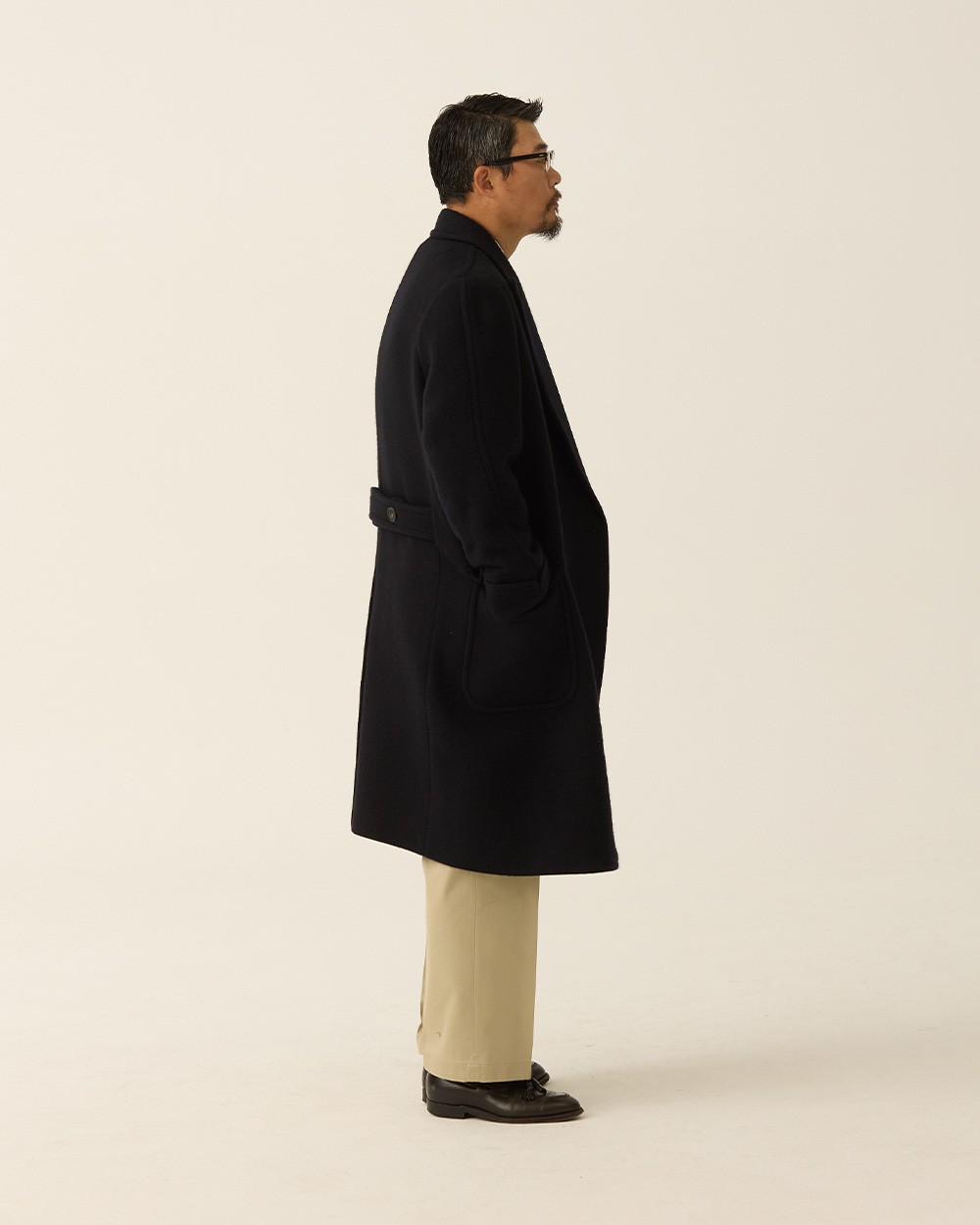 Side profile of a man in a navy polo coat buttoned, with khaki trousers and black loafers, showing the set-in sleeve and the half belt