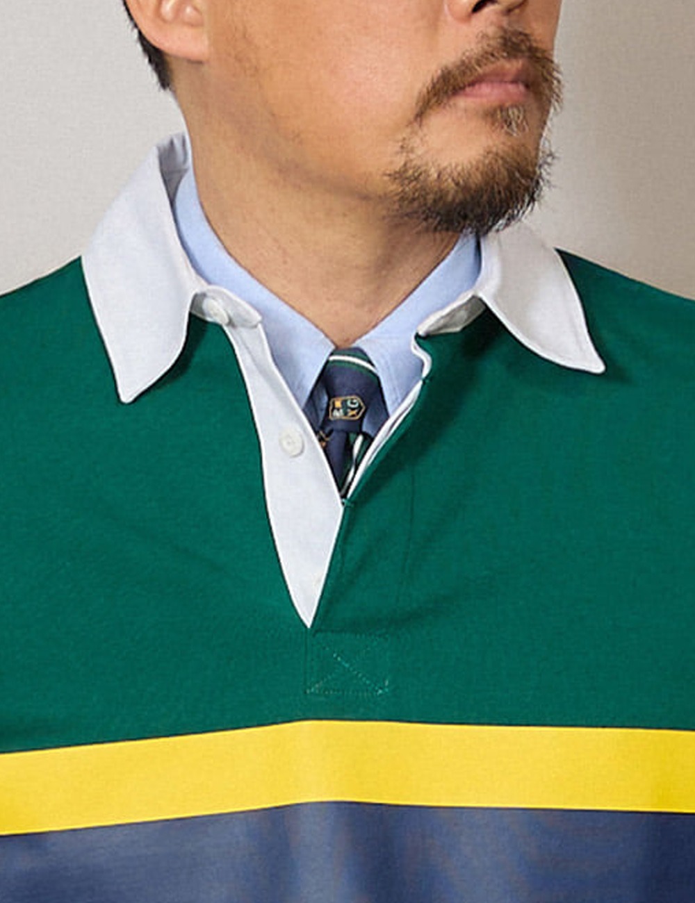 Close-up of a green rugby shirt's white collar lying over a light blue oxford shirt collar, with a navy club tie knot showing in the placket opening