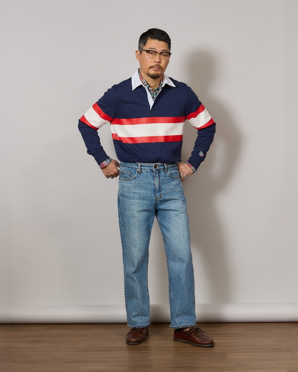 Man in a navy rugby shirt with a red and white chest band worn over a madras check shirt, light-wash jeans and brown penny loafers