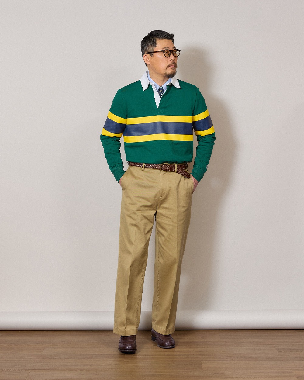 Man in a green rugby shirt worn over a light blue oxford button-down shirt and a navy club tie, tucked into khaki chinos with a braided leather belt and dark brown penny loafers