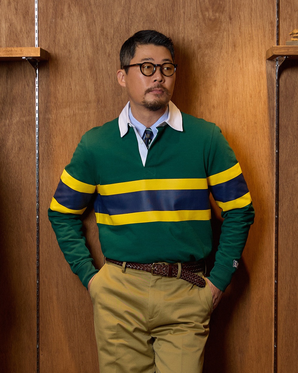Three-quarter view of a man in a green rugby shirt over an oxford shirt and tie, tucked into khaki chinos with a braided brown belt, standing against wood panelling