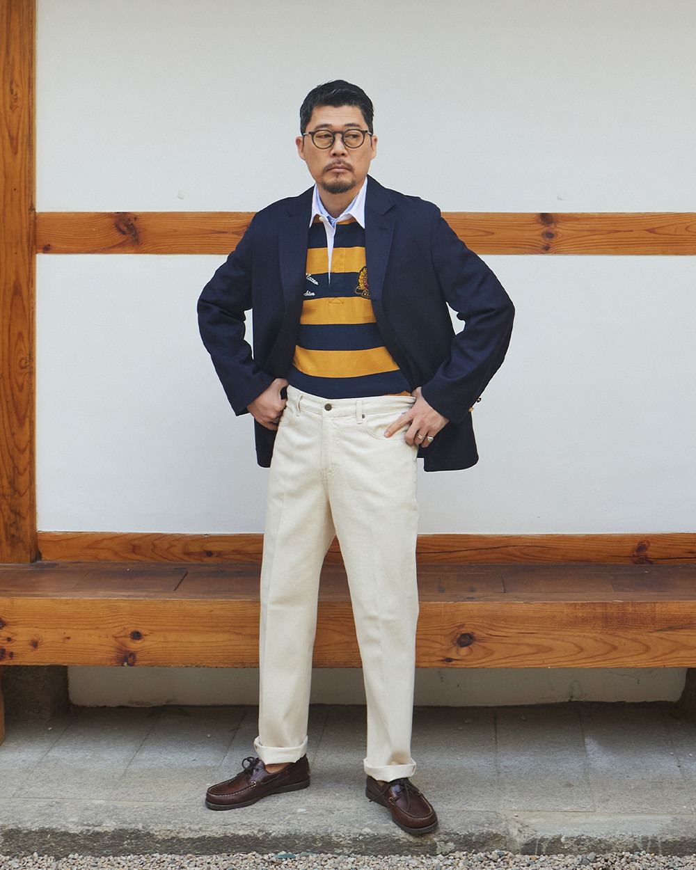 Man standing in front of a Korean hanok wearing a navy blazer over a yellow and navy hooped rugby shirt, white trousers and brown penny loafers