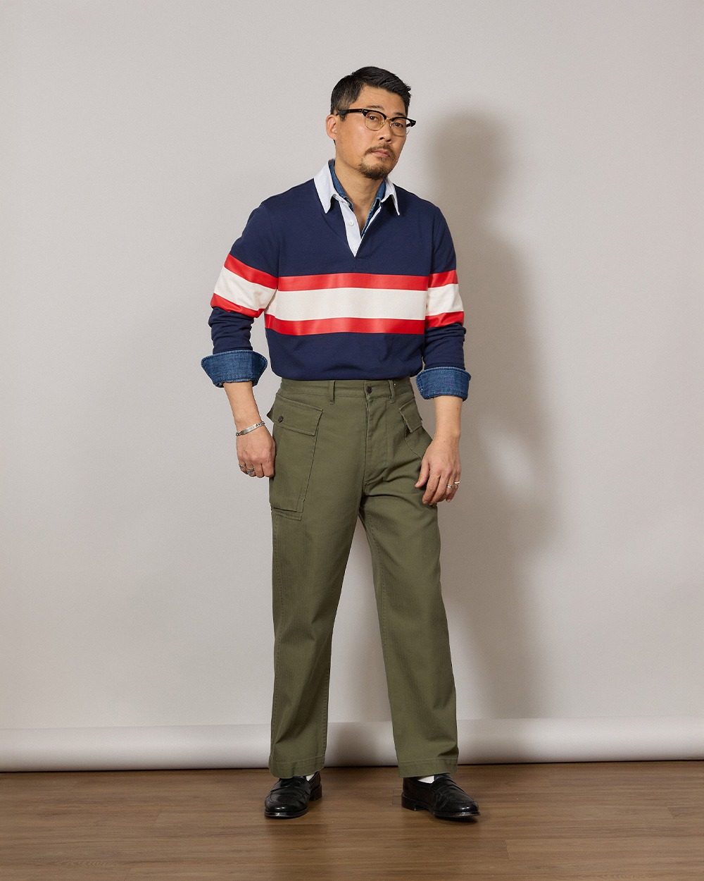 Man in a navy rugby shirt worn untucked over a chambray shirt, with the chambray cuffs turned back over the rugby cuffs, olive fatigue trousers and black loafers
